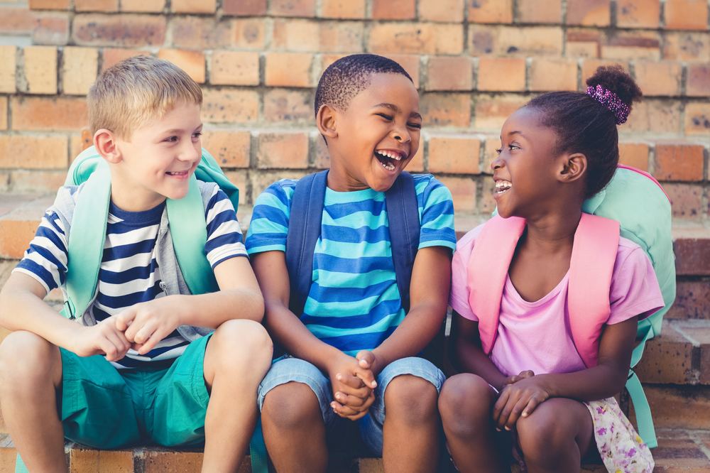 Three young students sitting together on brick steps outside school, laughing, with backpacks on.